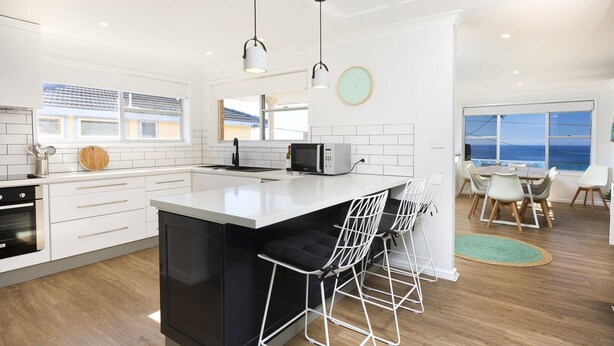 A modern kitchen with white cabinetry, a central island, bar stools, and a view of the ocean through large windows. - Aqua Palm with Sweeping Views of On Mile (Forster)