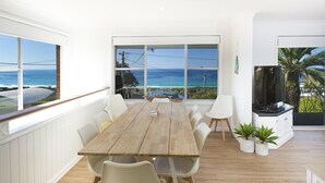 A dining area with a wooden table and white chairs, large windows with a view of the ocean, and a TV on a stand. - Aqua Palm with Sweeping Views of On Mile (Forster)