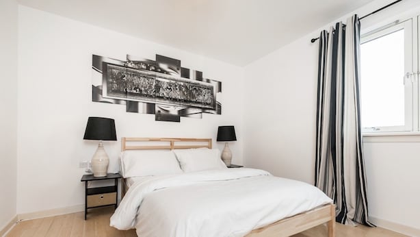 A bedroom with a wooden bed, two bedside tables with lamps, a large abstract artwork above the bed, and a window with curtains. - Stylish 2 Bedroom Apartment Aberdeen City Centre (Aberdeen)