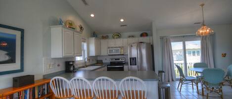 A kitchen with white cabinets, a central island with bar stools, a dining area with wicker chairs, and a view through a glass door.