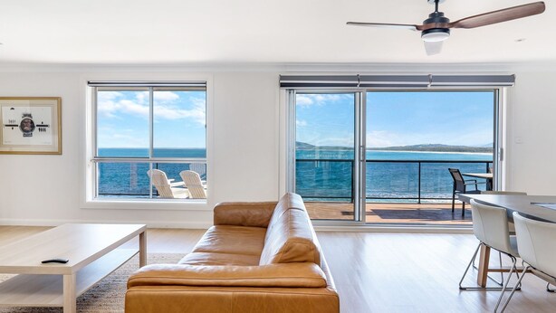 A modern living room with a leather sofa, a dining table, and chairs. Large windows offer a view of the ocean and a deck area. - Amaroo - Uninterrupted 180 degree water views (Gerroa)