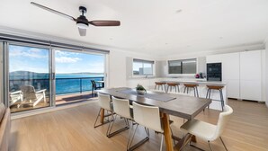 A modern kitchen with white cabinets, a dining area with a large table and chairs, and a view of the sea through large sliding doors. - Amaroo - Uninterrupted 180 degree water views (Gerroa)