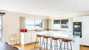 A modern kitchen with white cabinetry, a large island with stools, a built-in oven, and a view of the outdoors. - Amaroo - Uninterrupted 180 degree water views (Gerroa)