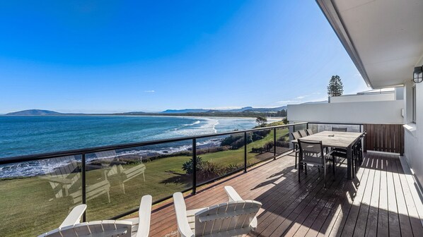 A balcony with a wooden deck, a table, and chairs overlooking a coastal view. - Amaroo - Uninterrupted 180 degree water views (Gerroa)