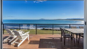 A deck with wooden flooring, white lounge chairs, and a table with chairs, overlooking a coastal view with the ocean and distant land. - Amaroo - Uninterrupted 180 degree water views (Gerroa)