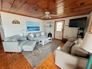 Ein Wohnzimmer mit grauem Ecksofa, einem weißen Couchtisch, einem grauen Teppich, einem Deckenventilator, einem Flachbildfernseher und einer Holzbalkendecke.