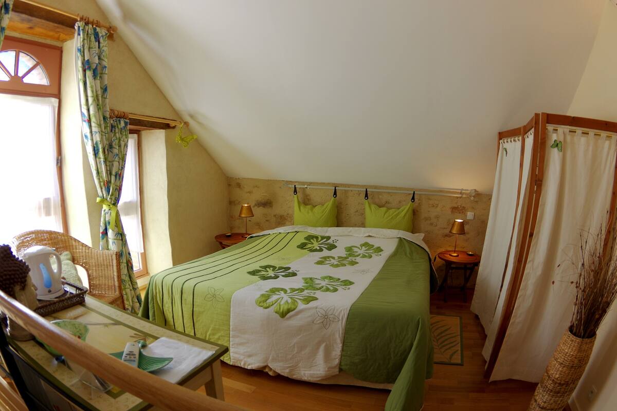 A bedroom with a sloped ceiling, a bed with green bedding, a wicker chair, a table with a kettle, and a lamp on a bedside table.
