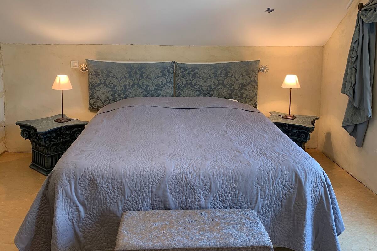 A bedroom with a large bed, two bedside tables with lamps, a bench, patterned wallpaper, and a curtain.