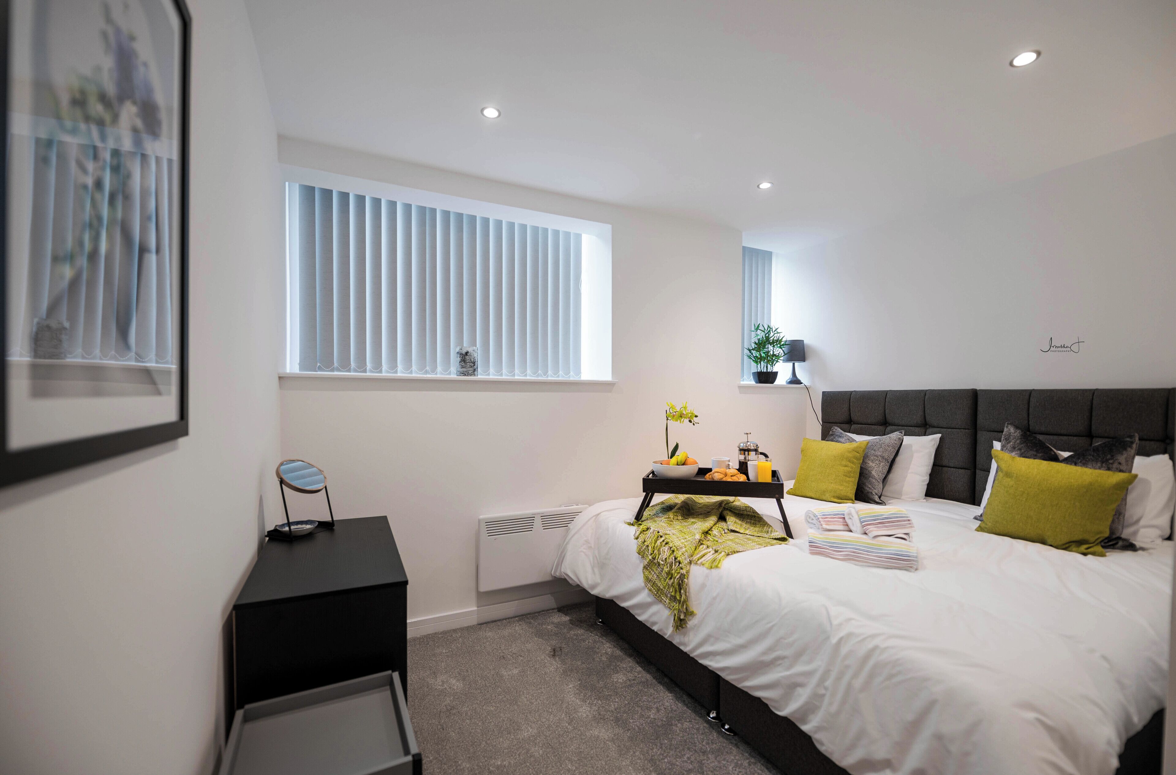 A modern bedroom with a large bed, bedside tables, a window with blinds, and a small plant.