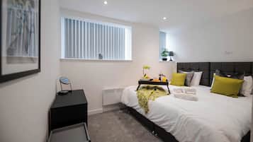 A modern bedroom with a large bed, bedside tables, a window with blinds, and a small plant.