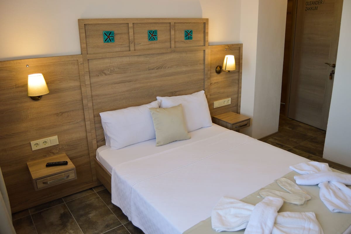 A hotel room with a wooden headboard, white bedding, a bedside table with a remote control, and a bathroom visible through an open door.