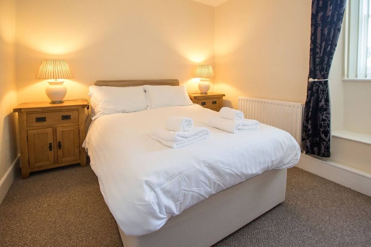 A neatly made bed with white linens and towels, two bedside tables with lamps, a radiator, and a window with curtains.