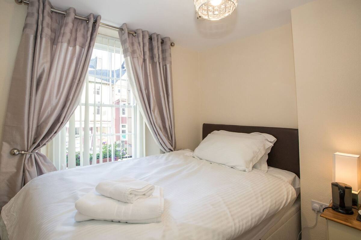 A neatly made bed with white linens and pillows, a bedside table with a lamp, and a window with curtains.