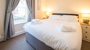 Uma cama bem arrumada com roupa de cama e toalhas brancas, uma mesa de cabeceira com abajur e uma janela com cortinas.
