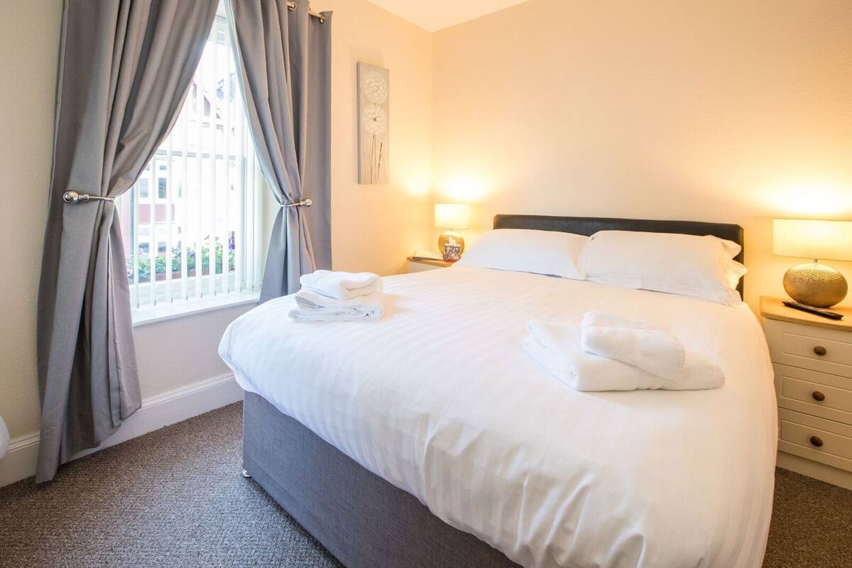 A neatly made bed with white linens and towels, a bedside table with a lamp, and a window with curtains.