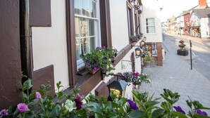 Um edifício com fachada branca, detalhes em marrom e várias janelas. Há caixas de flores com flores roxas no parapeito.