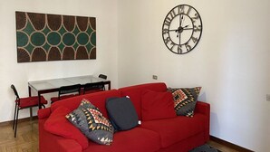 A living room with a red sofa, patterned cushions, a wall clock, and a dining table with chairs.