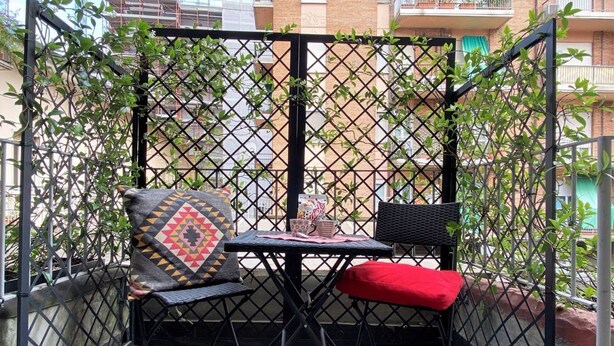 A small outdoor seating area with a table and two chairs, enclosed by a black metal fence with climbing plants.