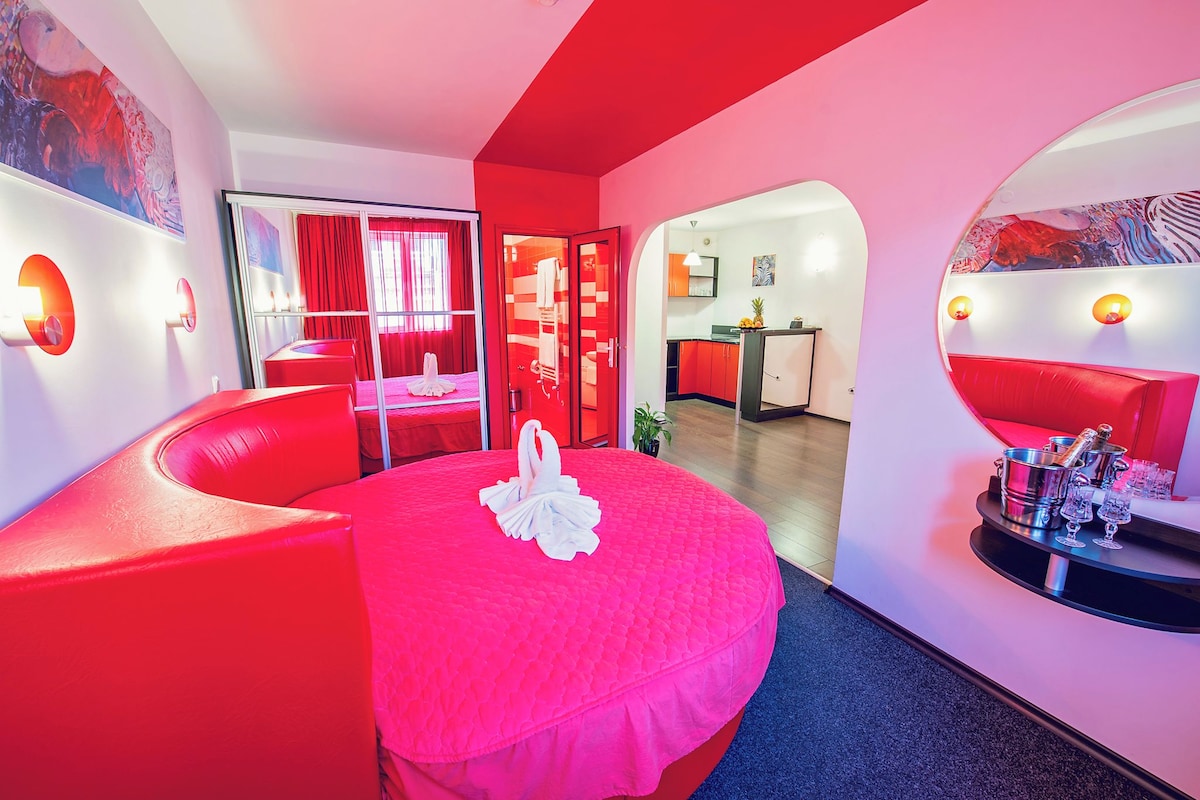 Una habitación de hotel moderna con una decoración roja, que incluye una cama redonda con adorno de cisne blanco, un sofá rojo y un minibar con champán.