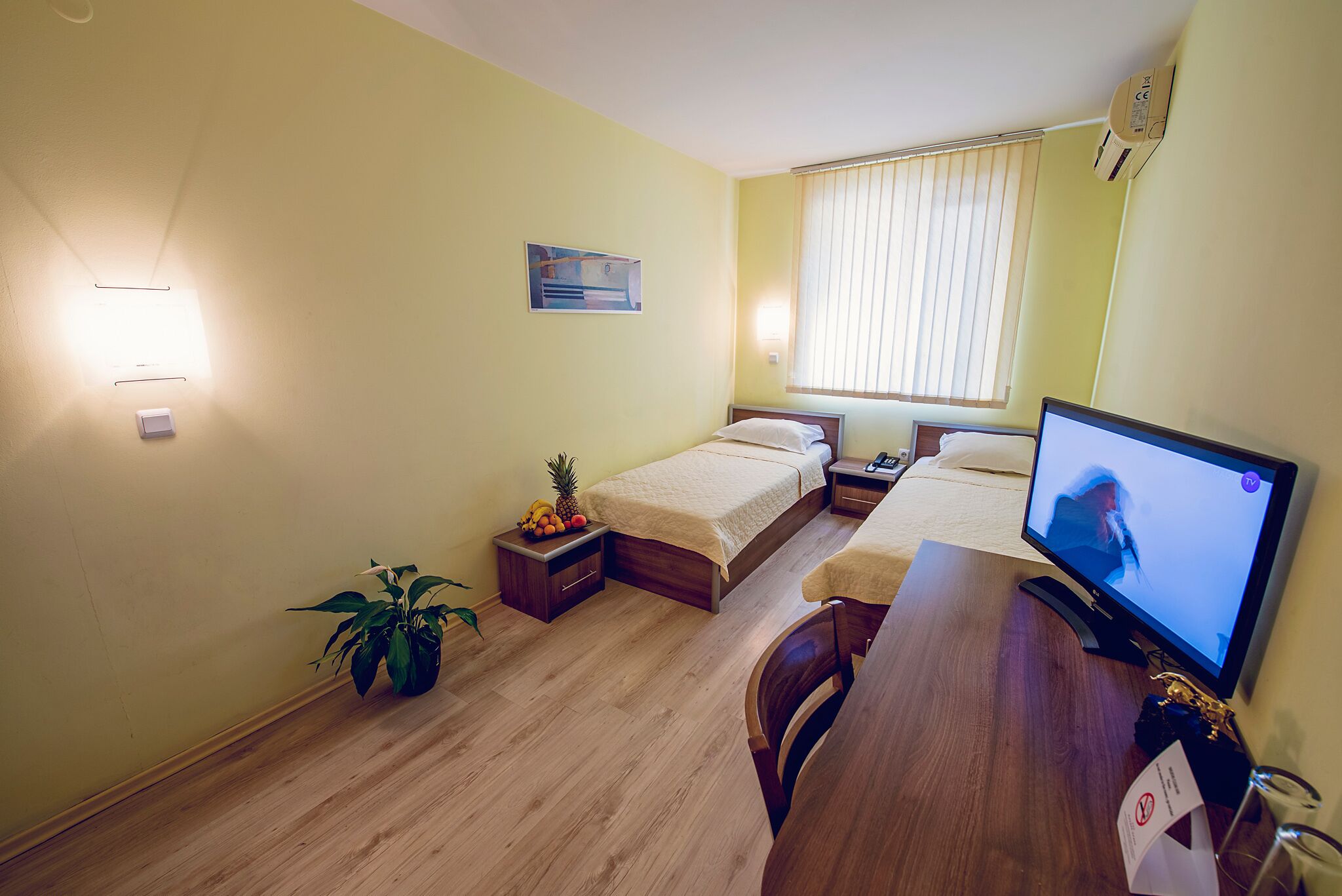 A small, well-lit room with two beds, a desk with a computer, a potted plant, and a window with blinds.