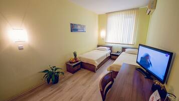 A small, well-lit room with two beds, a desk with a computer, a potted plant, and a window with blinds.