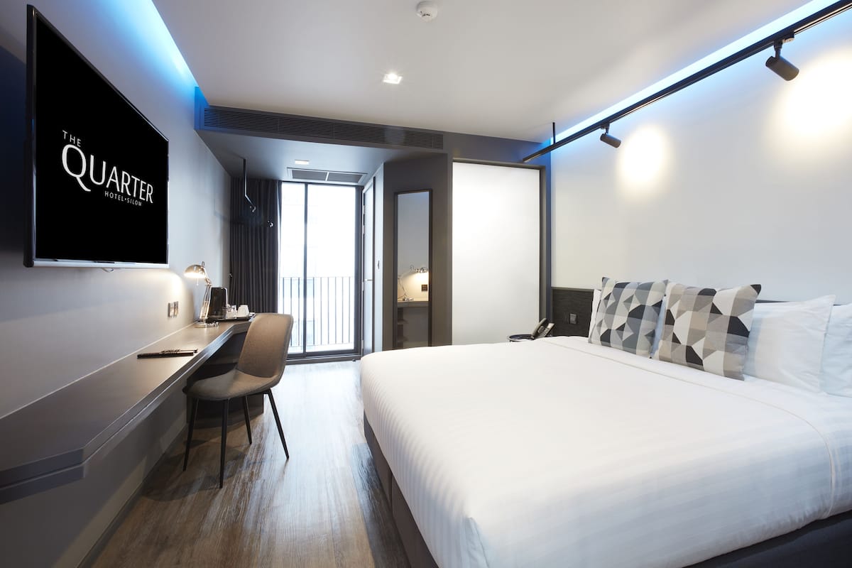 A modern hotel room with a large bed, a desk with a chair, a flat-screen TV mounted on the wall, and a balcony with a view.