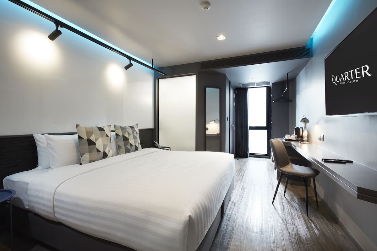 A modern hotel room with a large bed, a desk, and a chair. The room has a minimalist design with a large mirror and a sign that reads "QUARTER HOTEL SILOM".