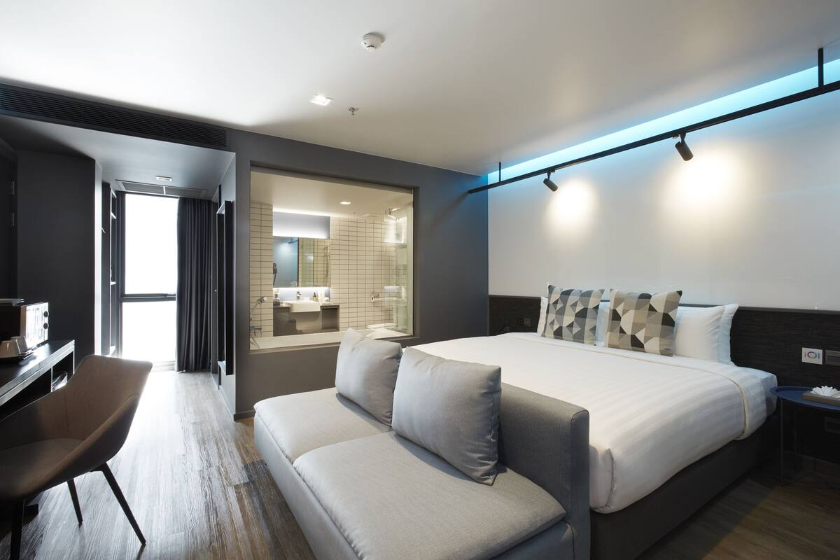 A modern hotel room with a large bed, a sofa, a desk, and a bathroom visible through an open door.
