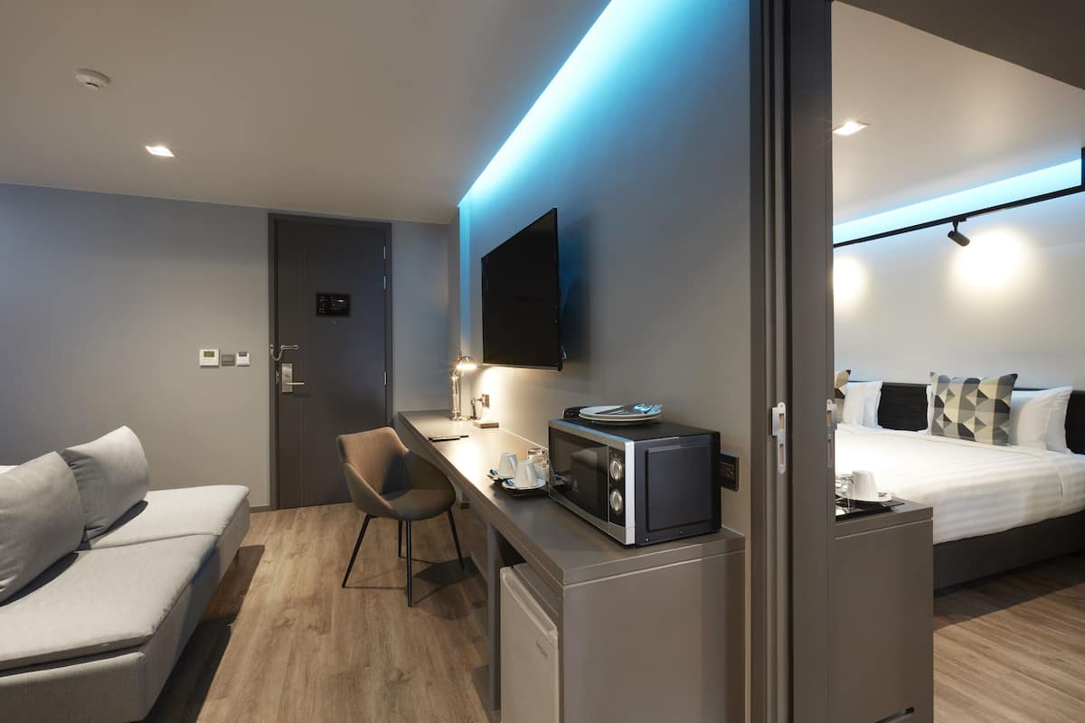A modern hotel room with a bed, a desk with a microwave, a chair, and a TV mounted on the wall.