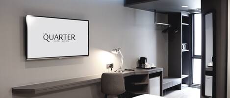 A modern hotel room with a bed, a desk with a chair, a desk lamp, a coffee maker, and a large framed sign on the wall.