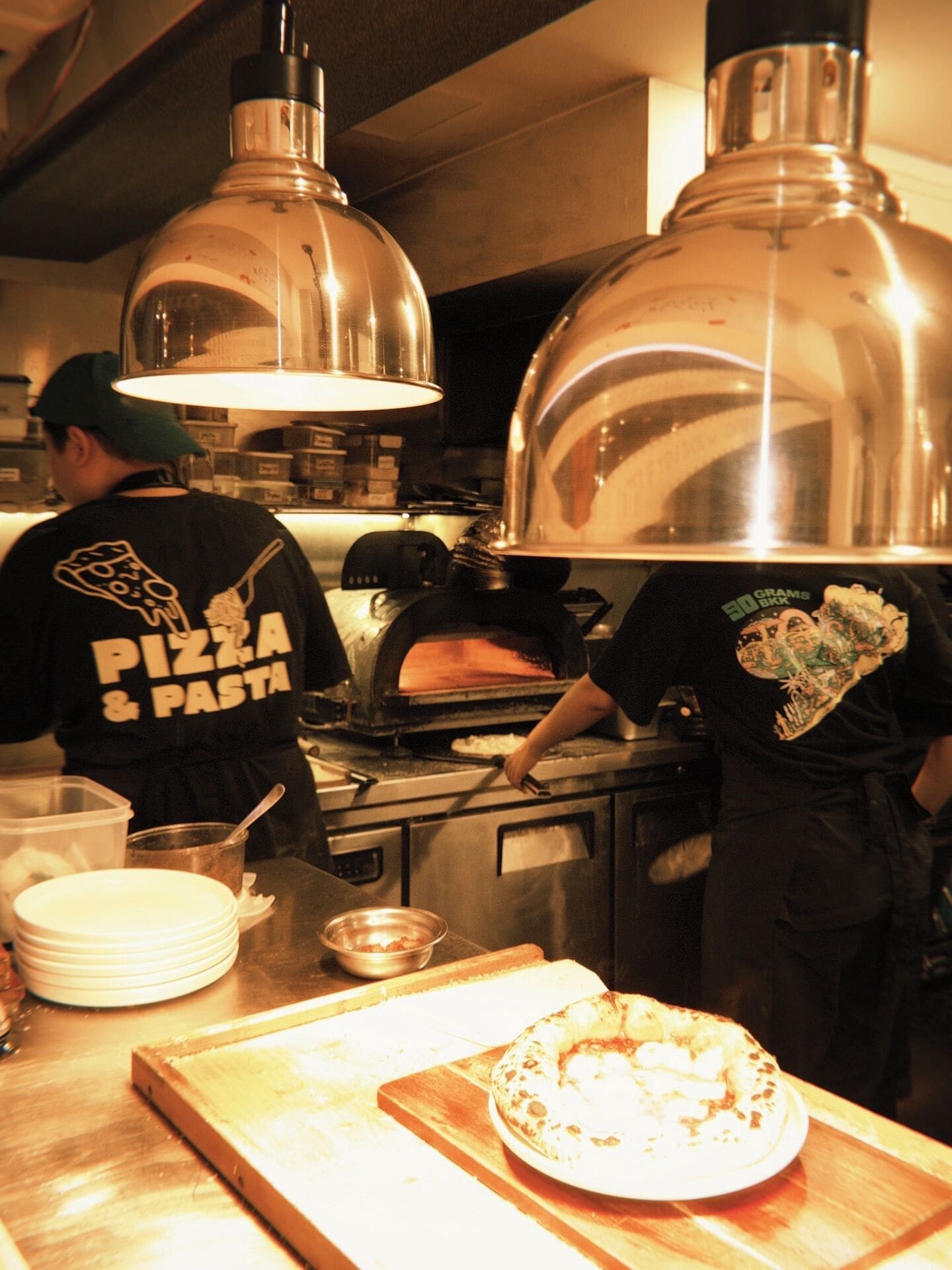 Una cocina con dos hornos, un horno de pizza, y una persona que lleva una camiseta con la inscripción 'Pizza & Pasta'.