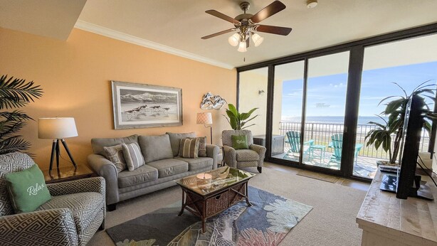 A spacious living room with a large window offering a view of the ocean, a ceiling fan, and a cozy seating area with patterned furniture.
