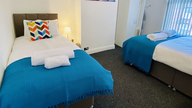Hotellihuoneessa on kaksi sänkyä, joissa kummassakin on sininen peitto ja valkoiset lakanat. Toisessa sängyssä on värikäs kuviollinen koristetyyny, ja seinällä sänkyjen yläpuolella on taulu.