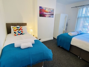 Een hotelkamer met twee bedden, elk voorzien van een blauwe sprei en witte lakens. Op een van de bedden ligt een kleurrijk kussen met een zigzaggemotief, en erboven hangt een schilderij aan de muur.