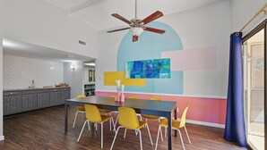 A modern kitchen with a dining area, featuring a large island with chairs, a ceiling fan, and a colorful abstract painting on the wall.