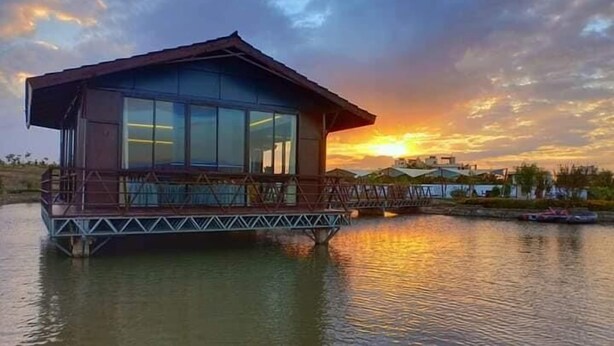 A floating building with a glass facade, a deck, and a metal railing, situated over water with a sunset in the background. - Storms Resort (Nanyuki)