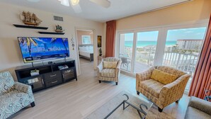 A living room with a TV, a sofa, a patterned armchair, and a balcony with a view. - Seacrest 706 is a 2 BR Gulf Front on Okaloosa Island by RedAwning (Fort Walton Beach)