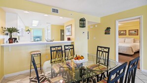 A dining area with a glass table and chairs, a kitchen in the background, and a bedroom visible through an open door.