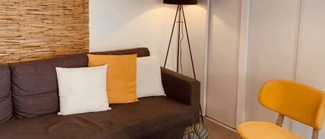 Een moderne woonkamer met een donkerbruine bank, een gele fauteuil, een witte salontafel, een vloerlamp en een hangende pendellamp.