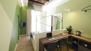 Habitación pequeña con cama, escritorio con una computadora portátil, dos sillas, una mesita con una planta y una ventana con cortinas.