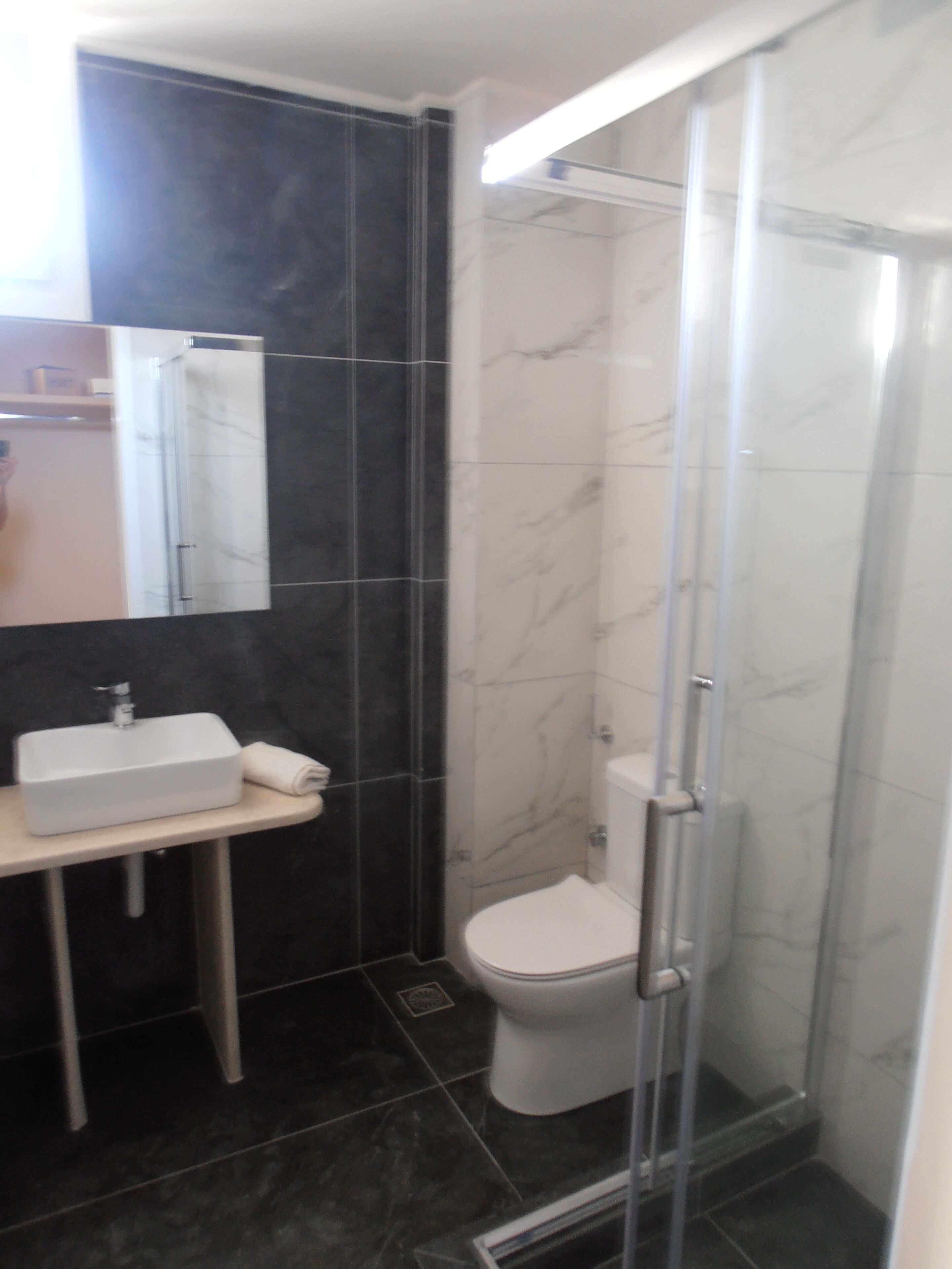 A bathroom with a white sink, dark tiled walls, a white toilet, and a glass shower enclosure.