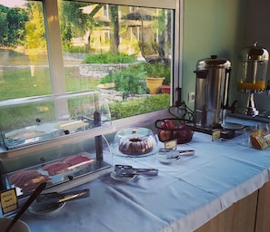 A breakfast buffet with various food items and a coffee machine, set against a window with a view of greenery and buildings. - Frosini Gardens (Corfu)
