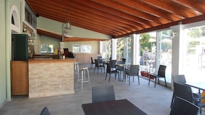 An outdoor cafe area with wooden ceiling beams, a bar counter, and tables and chairs. - Frosini Gardens (Corfu)