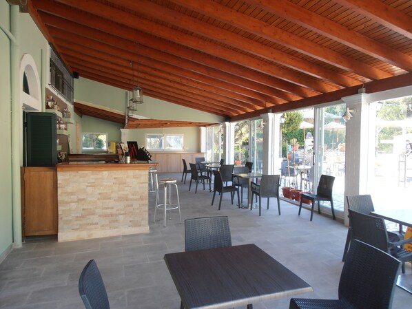 An outdoor cafe area with wooden ceiling beams, a bar counter, and tables and chairs.
