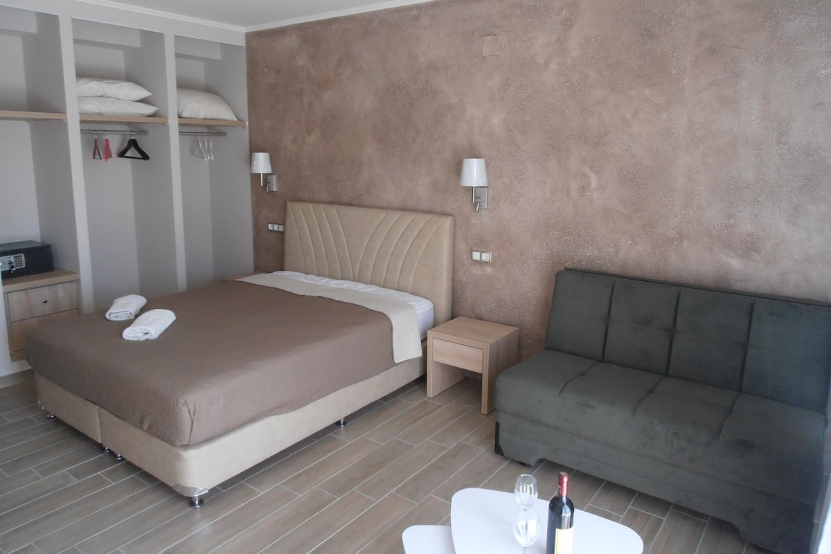 A modern hotel room with a large bed, a grey sofa, a small table with a bottle and glasses, and a wardrobe with hangers.