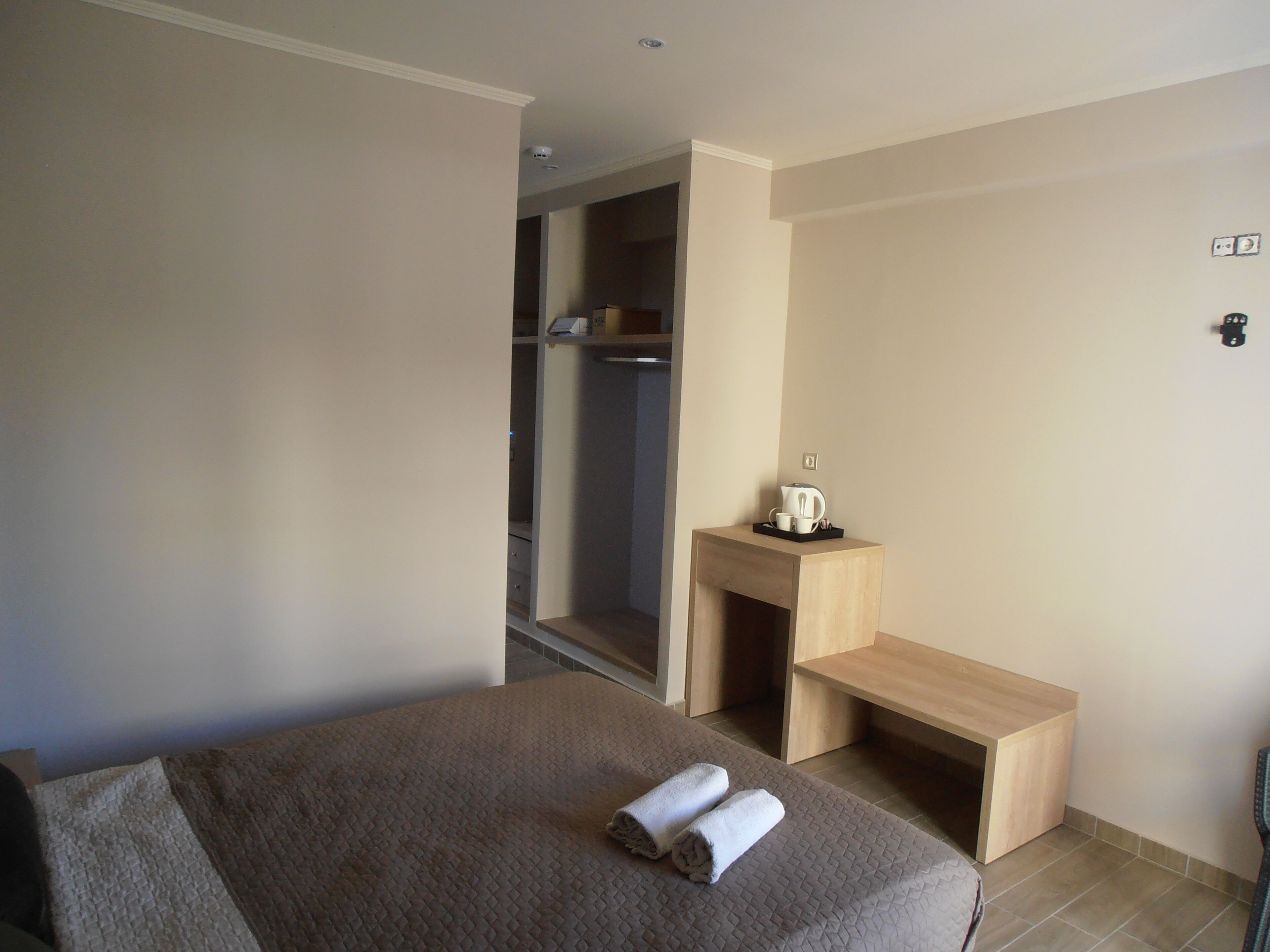 A hotel room with a bed, a wooden nightstand, a small table with a kettle and cups, and a closet.