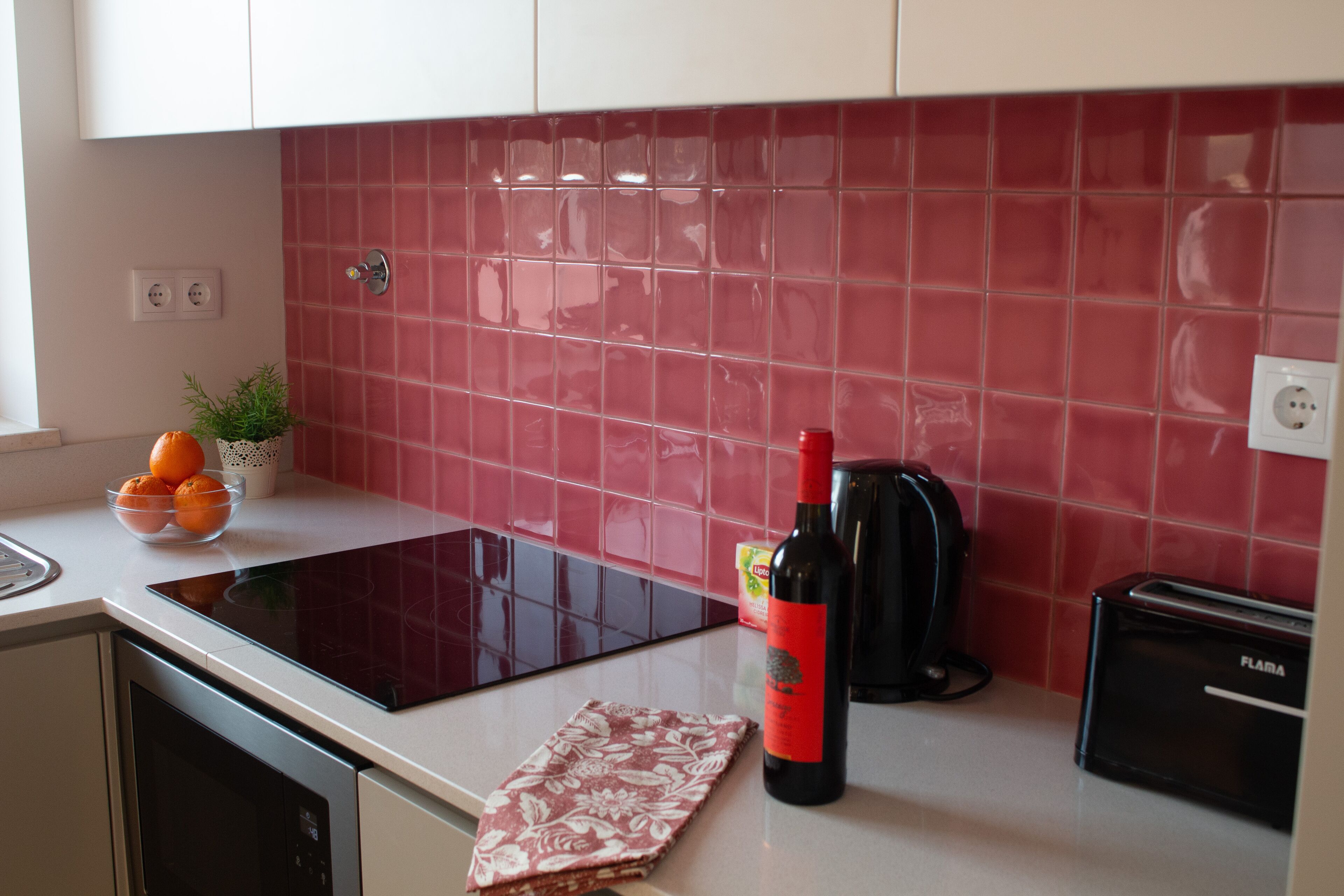 A kitchen with a red tiled backsplash, a black countertop, a wine bottle, a toaster, and a bowl of fruit.