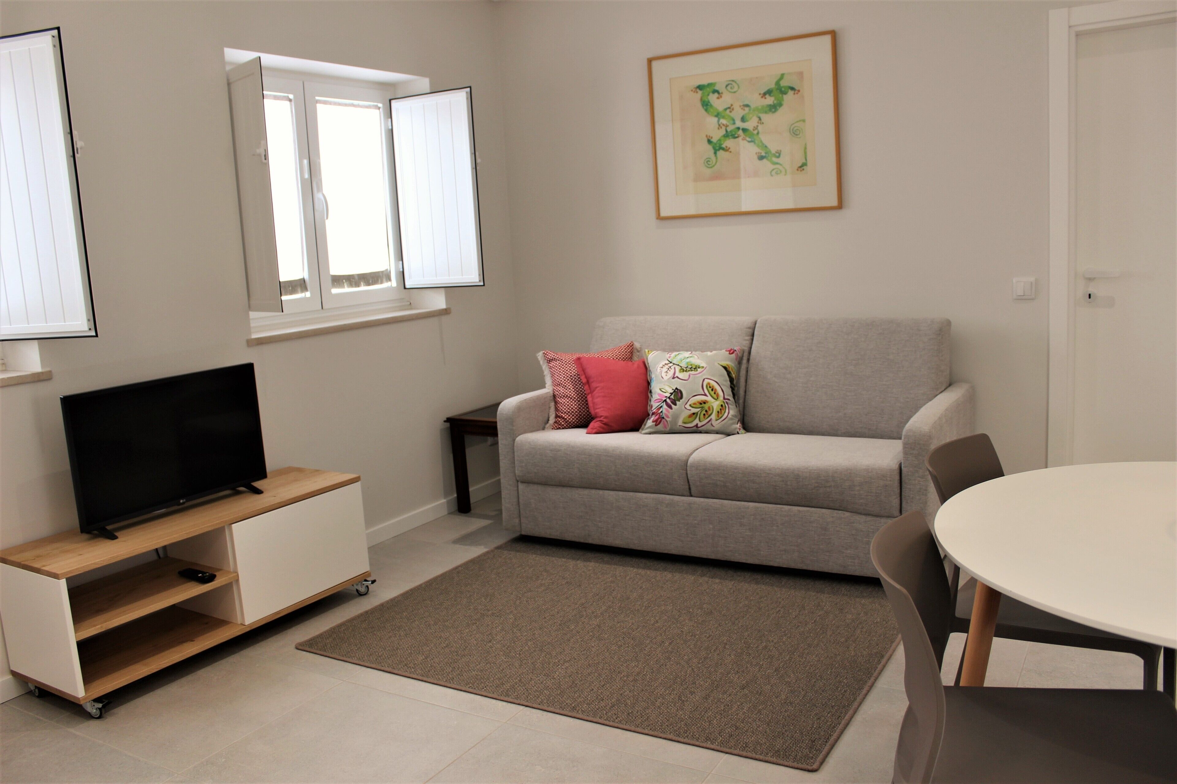 A living room with a sofa, a TV on a stand, a dining table, and a window with blinds.