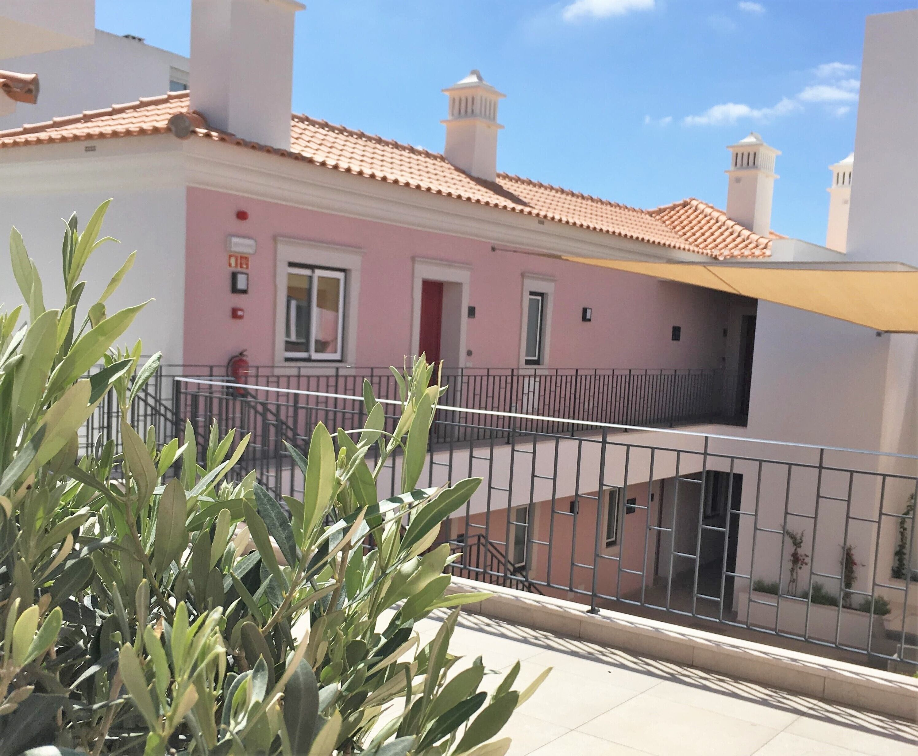 A pink building with a tiled roof, a balcony with a metal railing, and a plant in the foreground.