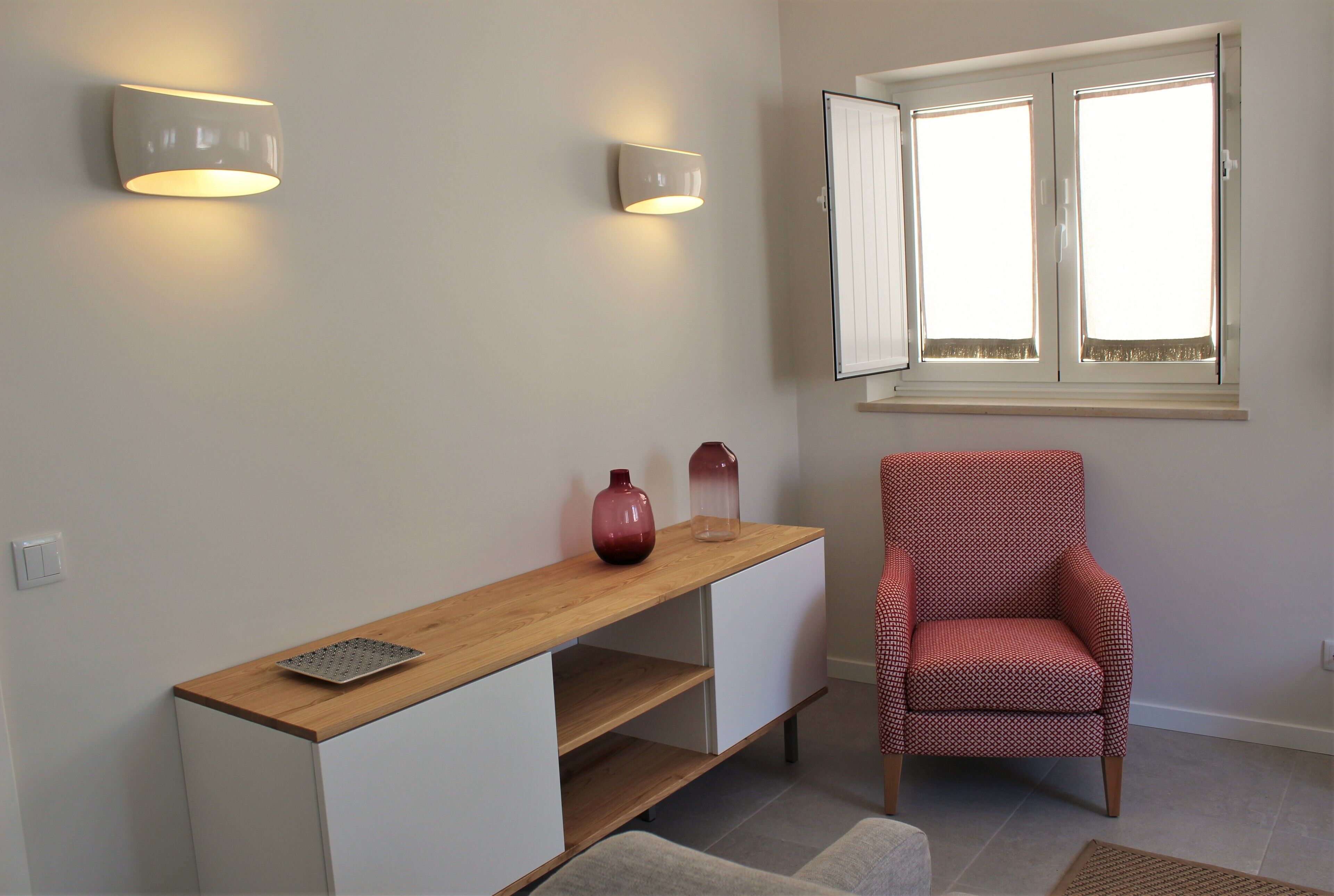 Ein Zimmer mit einem Holztisch, zwei Wandleuchten, einem rot gemusterten Stuhl, einem Fenster mit Jalousien und einem kleinen Teppich auf dem Boden.